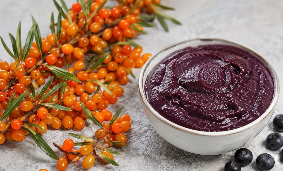 Fresh sea buckthorn berries beside a bowl of dark purple acai puree on a bright surface