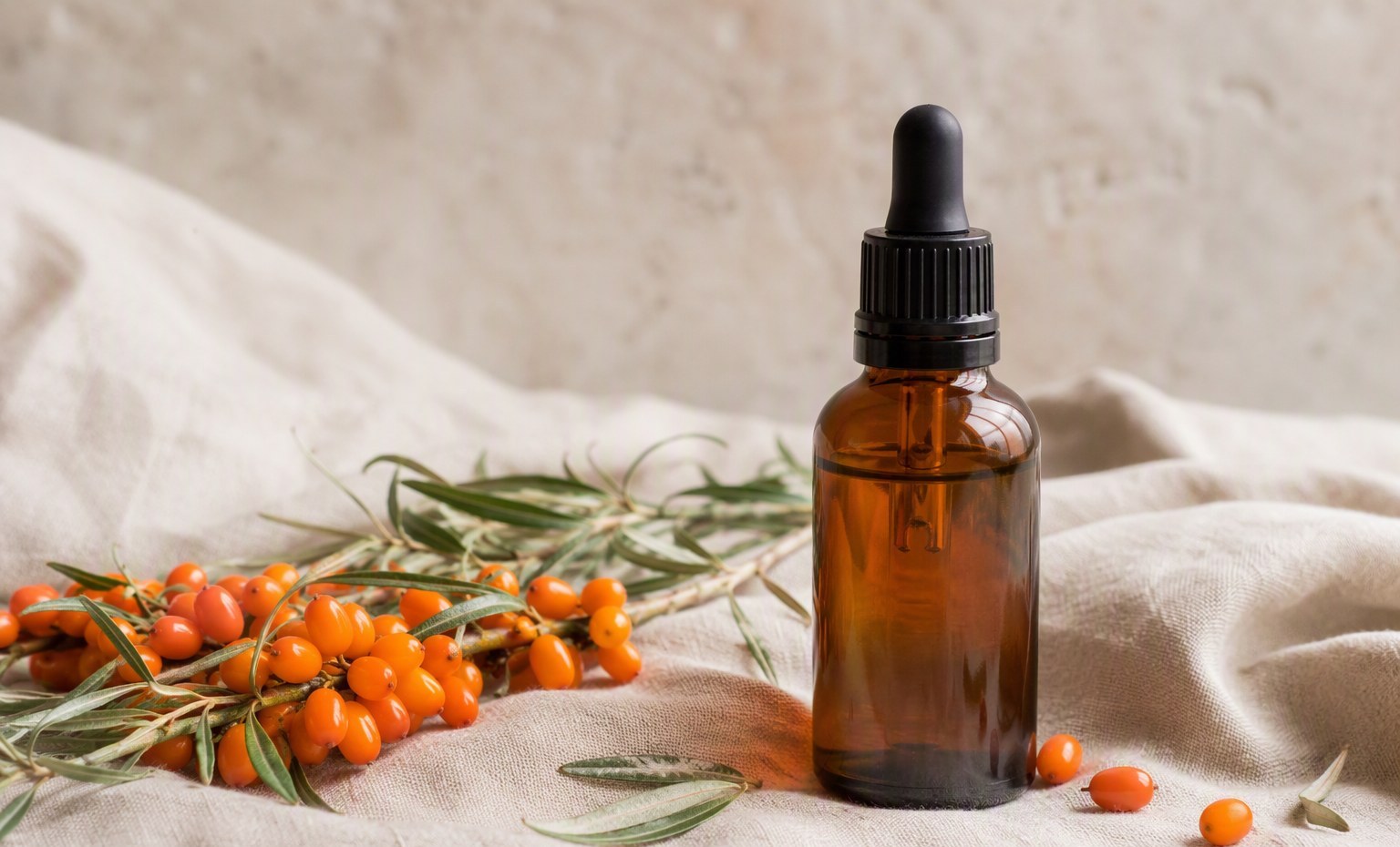 An unbranded amber sea buckthorn oil bottle on natural linen with fresh berries and leaves