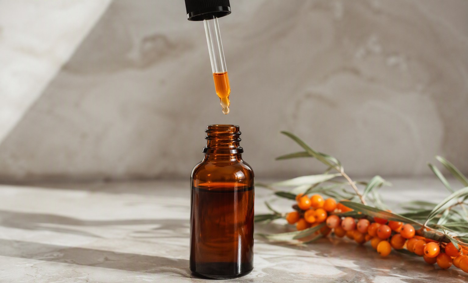A dropper releasing a drop of orange sea buckthorn oil into an open amber bottle on pale stone