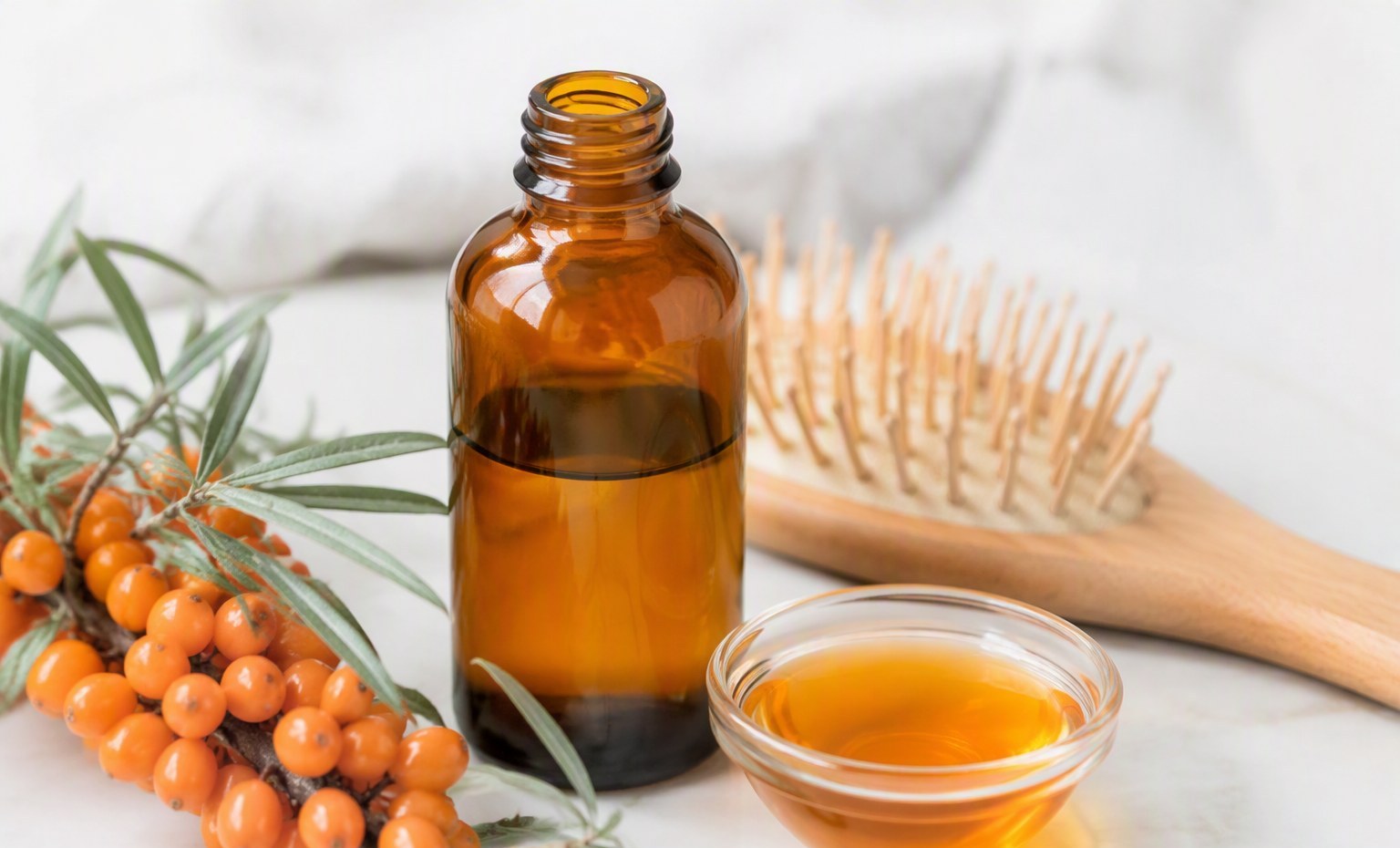 A sea buckthorn hair oil bottle beside a wooden brush and a small dish of orange oil