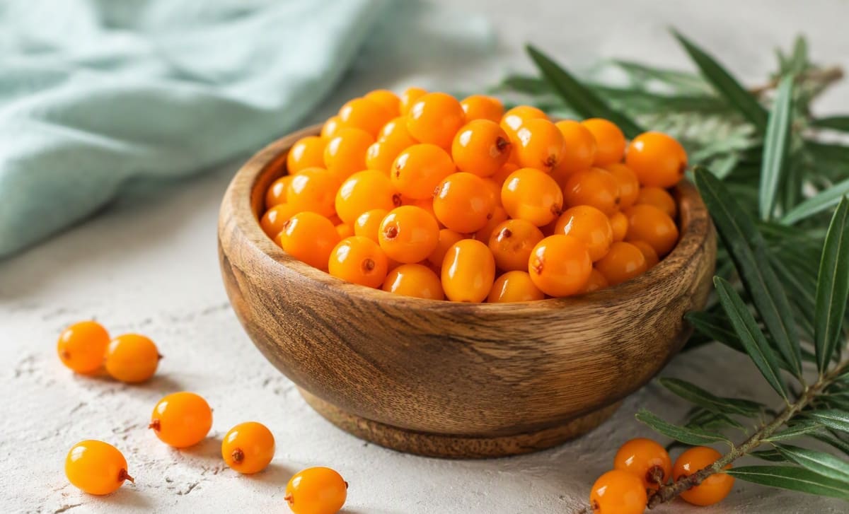 A wooden bowl of fresh sea buckthorn berries with scattered berries and leaves on a bright table