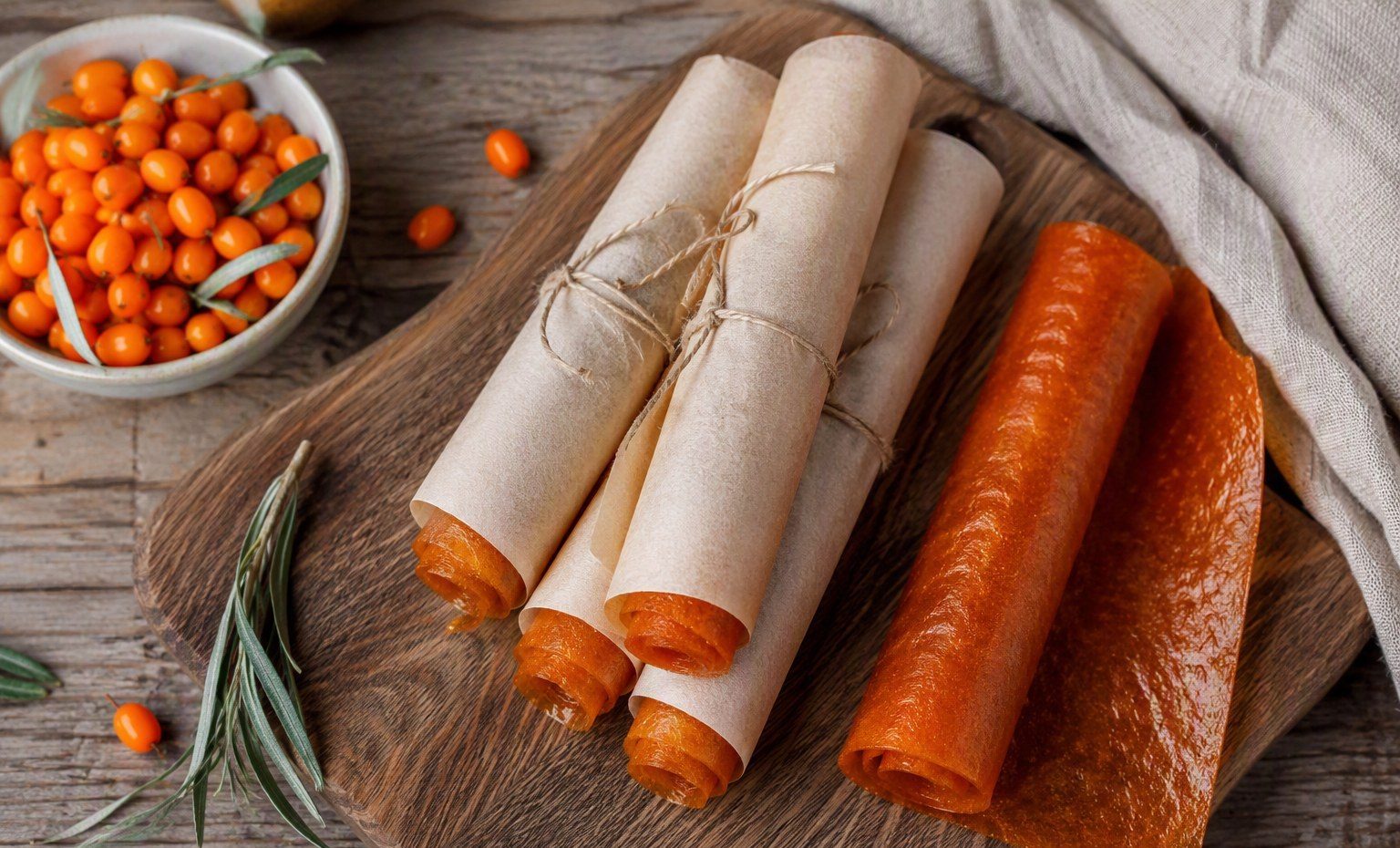 Orange sea buckthorn fruit leather rolled in parchment on a wooden board beside a bowl of berries