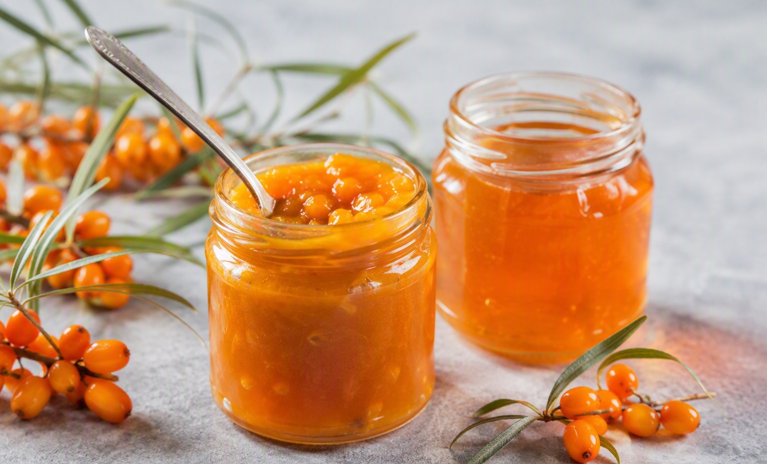 Two open jars of bright orange sea buckthorn jam with a spoon and fresh berries