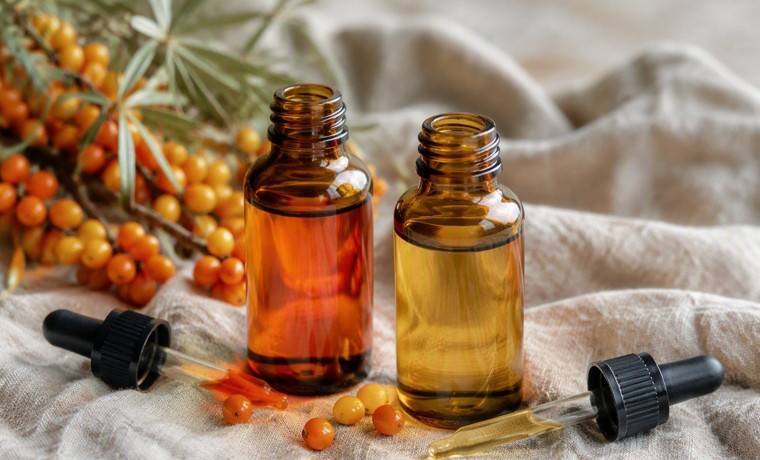 Two amber bottles of sea buckthorn oil, deep orange berry oil and pale seed oil, with droppers on linen