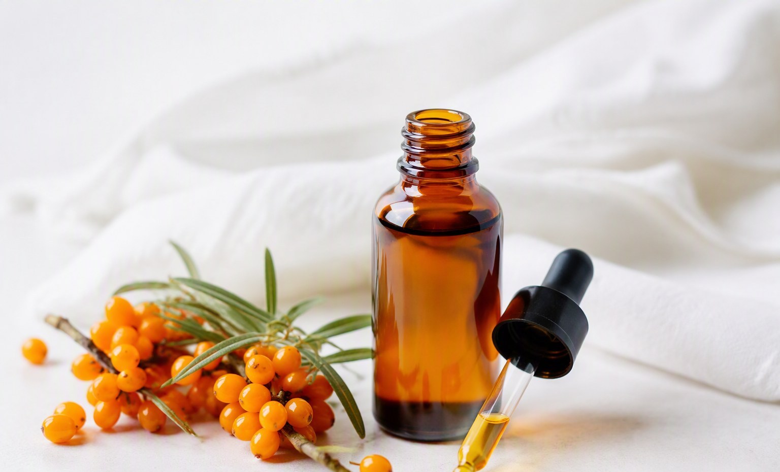 An amber dropper bottle of sea buckthorn oil with fresh berries on a soft white background