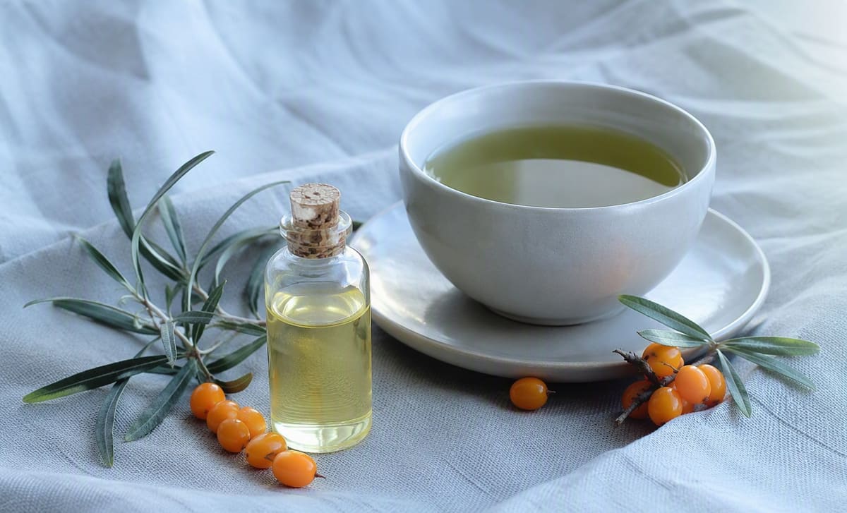 A cup of green tea beside a small bottle of pale sea buckthorn seed oil on cool blue linen
